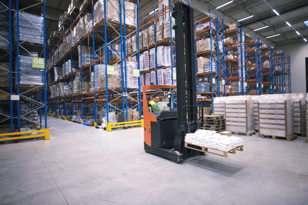 Worker handling material by operating forklift machine and relocating goods in large warehouse center.